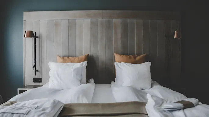 En pent oppredd seng med hvitt sengetøy og beige puter står under et trepanel, flankert av to veggmonterte kobberlamper, i en koselig, minimalistisk romsetting.