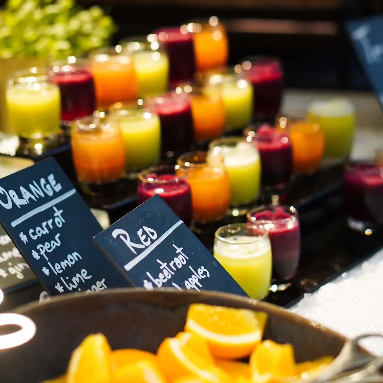 Fargerike juiceglass arrangert på et bord, omgitt av frisk frukt. Etiketter oppgir ingredienser: "Orange: gulrot, pære, sitron, lime, ingefær" og "Red: rødbet, epler". Sitroner og lime er synlige.