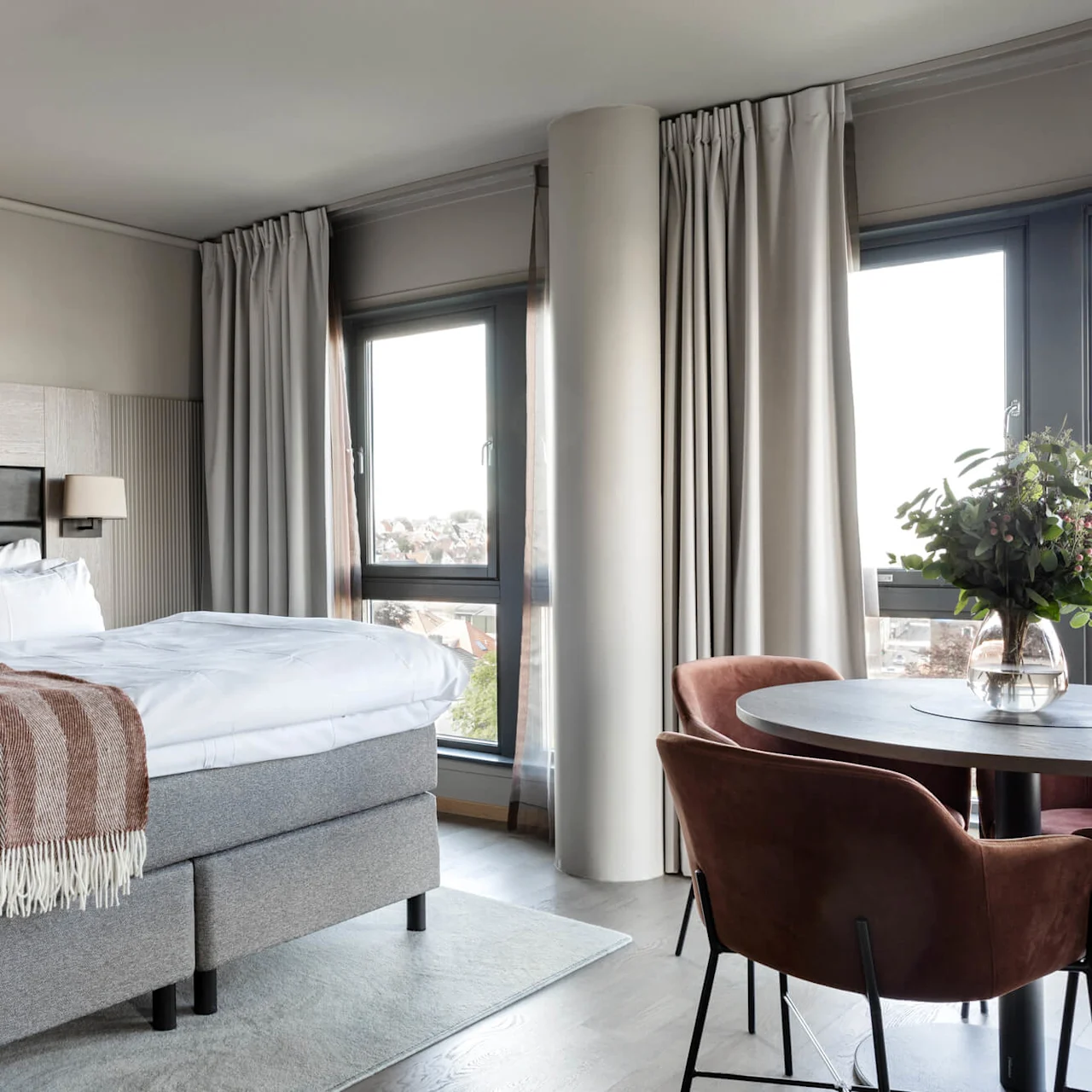 A neatly made bed with a striped throw lies beside a small round table with flowers. The room is bright with large windows and cream curtains.