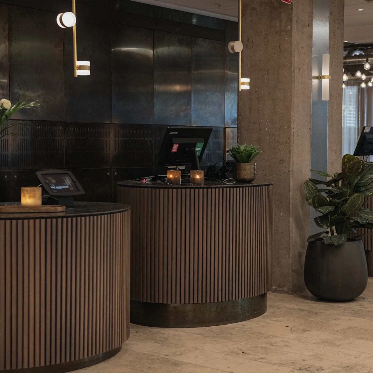 Three round wooden reception desks with computer monitors and decorative plants, positioned in a modern lobby featuring pendant lights, a concrete floor, and walls with a dark, metallic finish.