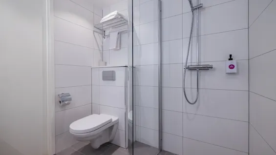 Toilet mounted on a white-tiled wall with a paper holder nearby; adjacent to a glass-enclosed shower area with silver fixtures. Minimalist bathroom with overhead shelf holding towels.