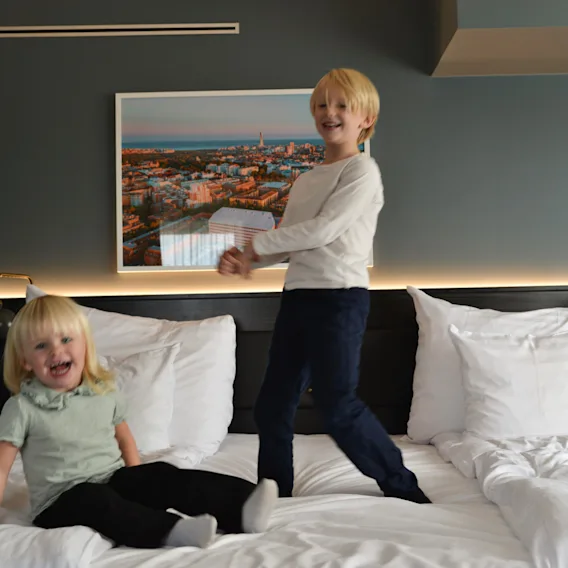 Two children jump and sit on a white hotel bed, with a city photo on the wall behind them and soft ambient lighting.