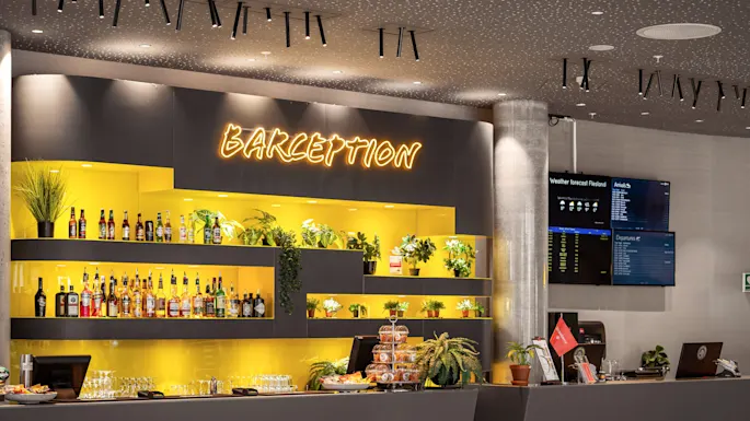 Bar with yellow-lit shelves displaying bottles and plants. Neon sign reads "BARCEPTION". Wall-mounted screens show "Weather forecast Flesland", "Arrivals", and "Departures". Snacks and glasses on the counter.