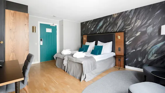 A tidy hotel room with twin beds, teal pillows, dark headboard, and feather-patterned wallpaper. A desk, chairs, and wood flooring enhance the space.