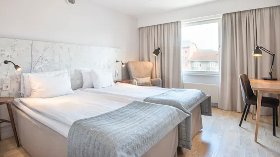 Två sängar med vita lakan och grå filtar är placerade i mitten av ett hotellrum, omgivna av sänglampor, en fåtölj och ett skrivbord. Ett fönster avslöjar byggnader utanför.