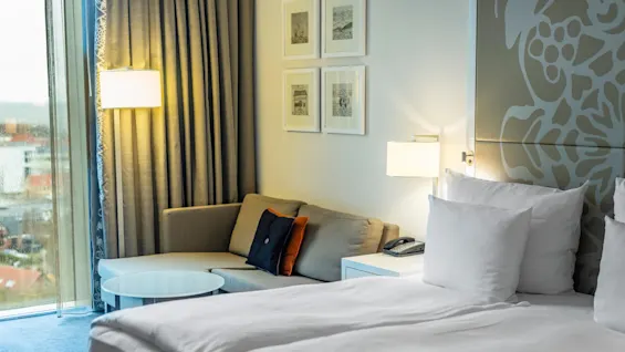 En prydligt bäddad säng med vita kuddar står i ett modernt hotellrum. Bredvid finns en soffa och ett glasbord vid ett fönster med tjocka gardiner, bredvid inramad väggkonst.