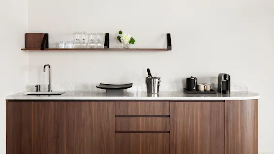 En köksbänk med en diskho har en kaffemaskin, koppar och en vinkylare; ovanför, en hylla visar glas och en vas med vita blommor mot en vit vägg.