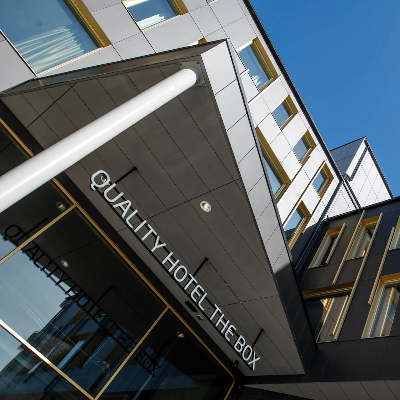 Facade of a modern building with large windows; entrance displays "QUALITY HOTEL THE BOX" in clear letters. The sky is bright blue, highlighting the structure's geometric design. 