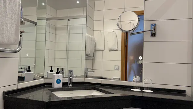 Et hvitt flislagt bad har en sort benkeplate med en vask, under sterkt lys. Speil og et håndklehenger pryder veggene. To glass og toalettsaker ligger ved siden av vasken.