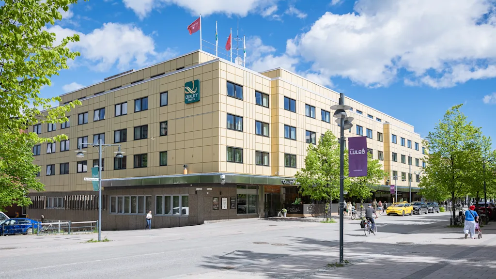 En stor hotellbyggnad med en grön logotyp under blå himmel, med människor som går och cyklar framför.