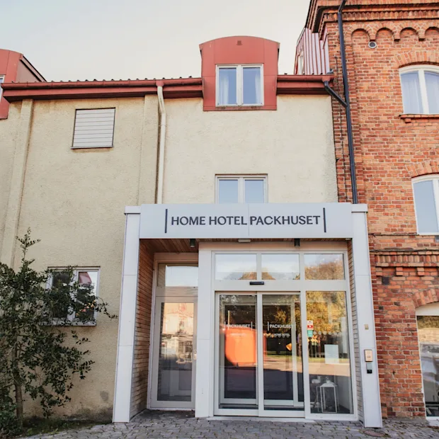 Indgang til en hotelbygning med et skilt, der læser HOME HOTEL PACKHUSET over de automatiske glasdøre.