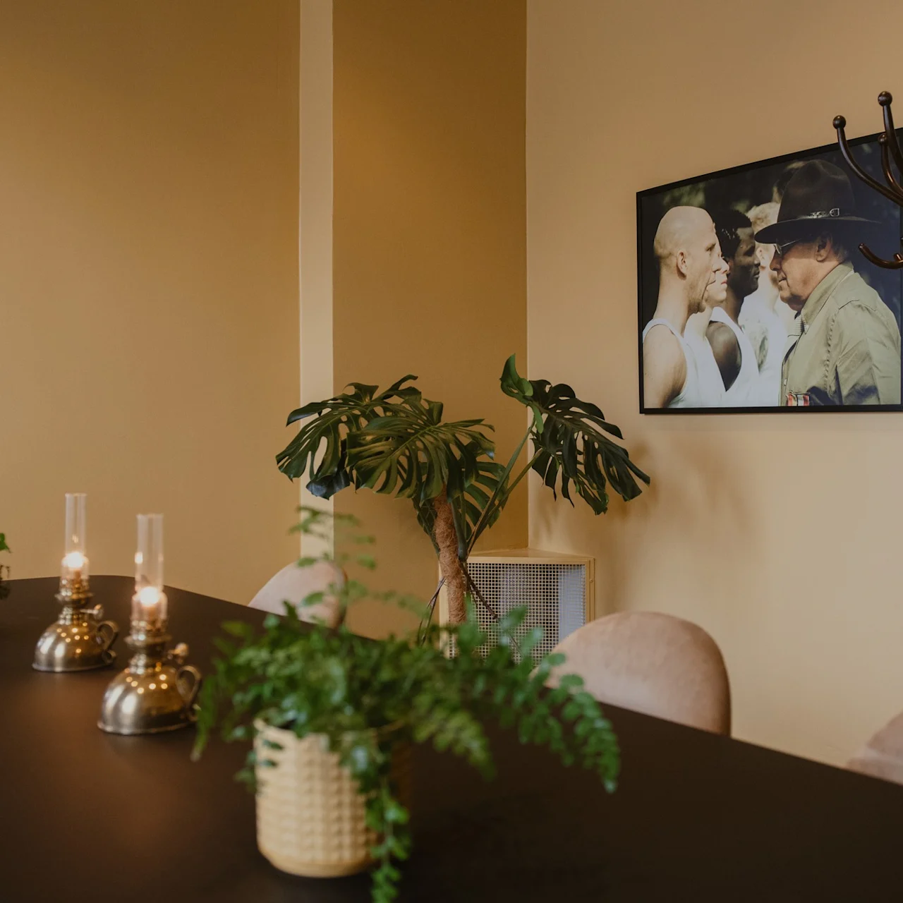 A black table holds two lit oil lamps and potted plants. Nearby, a framed photo hangs on a beige wall. A coat stand is placed beside the table.