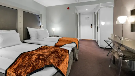 To pænt redte enkeltsenge med orange tæpper står ved grå vægge. Et skrivebord med en gennemsigtig stol og lampe står i hjørnet og skaber en ryddelig hotelfølelse.