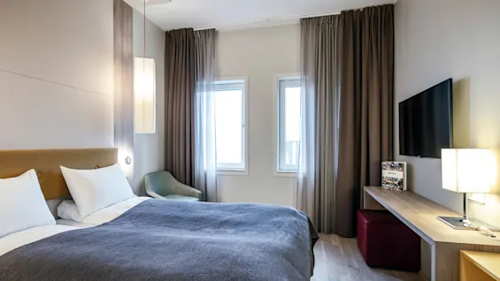 Et koselig hotellrom har en pent oppredd seng med et grått teppe ved siden av et lite bord med stol, en veggmontert TV og store vinduer med gardiner.