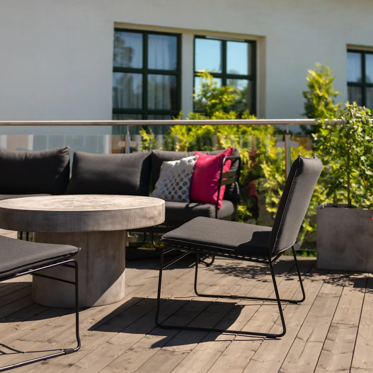 Outdoor seating set surrounds a round stone table on a wooden deck, with vibrant cushions and potted plants against a building backdrop.