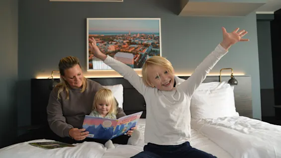 Et barn med hævede arme sidder glad på en hotelseng ved siden af en kvinde, der læser for et andet barn. Et bylandskab hænger på den mørke væg i baggrunden.