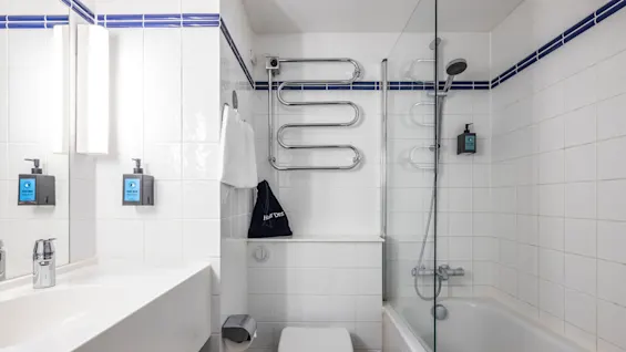 A modern bathroom with white tiles and blue accents. It features a sink with a mirror and integrated lighting, a wall-mounted toilet, and a bathtub with a shower. 