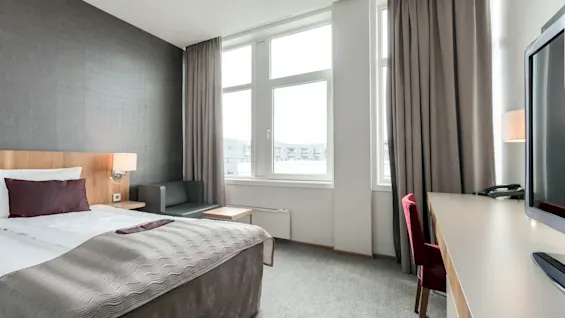 Siisti hotellihuone, jossa on huolellisesti pedattu sänky teksturoidun seinän edessä, sohva, työpöytä tuolin ja television kanssa suurten ikkunoiden vieressä.