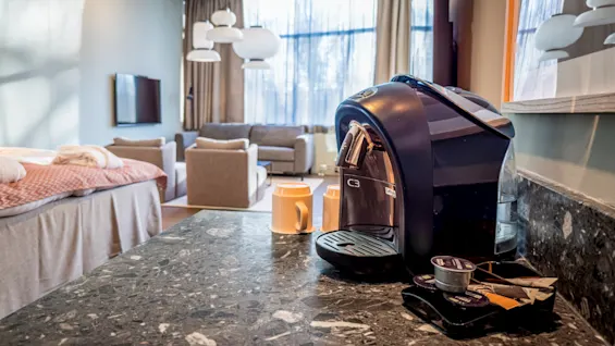 A black coffee machine sits quietly on a marble countertop, accompanied by mugs and capsules, in a cozy living room with a bed, couches, and large windows allowing natural light.