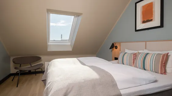 Ett ljust och inbjudande hotellrum med en bekväm säng, ett takfönster och en modern stol, perfekt för en avkopplande vistelse.