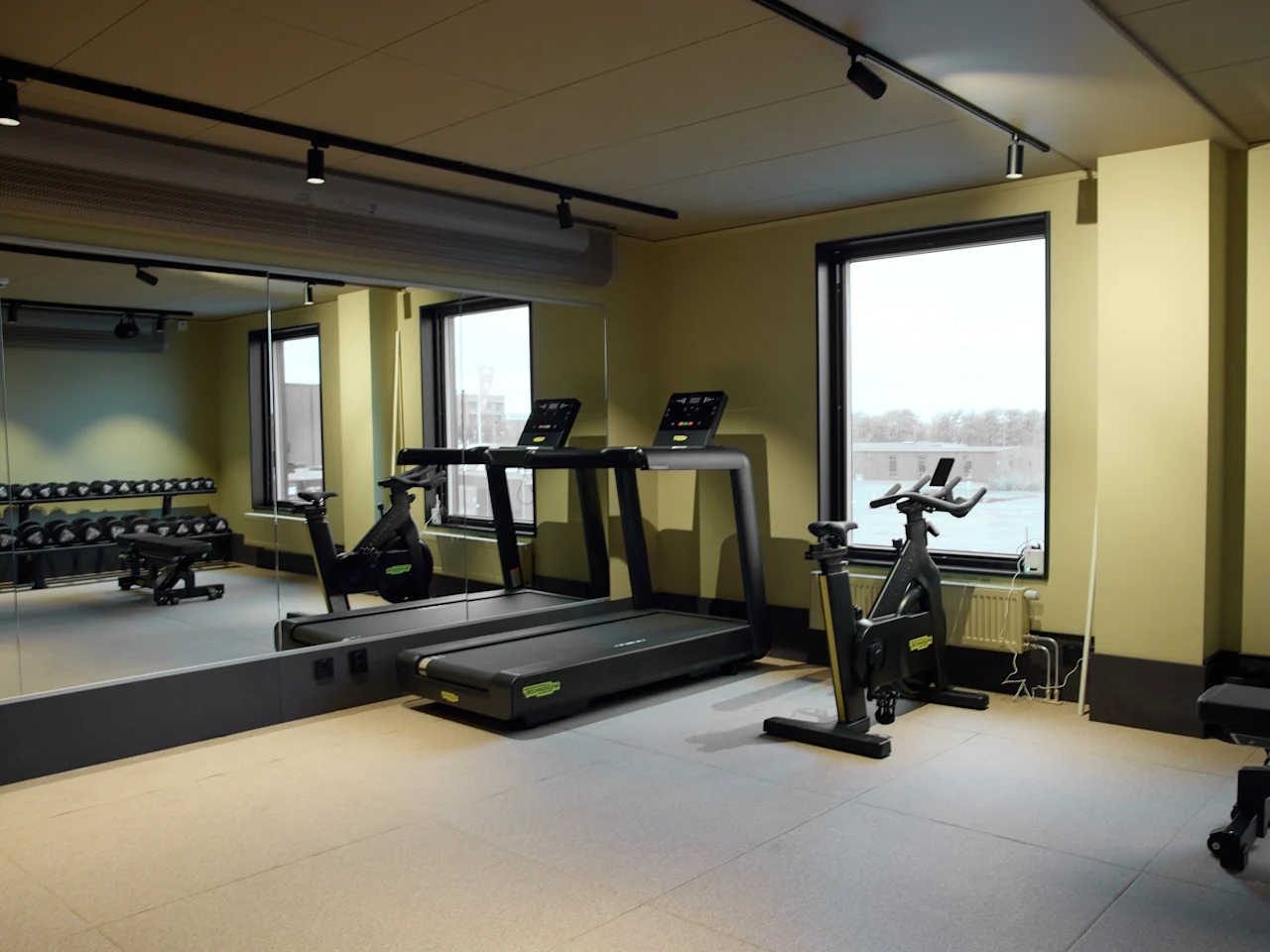Ett modernt gym med löpband, en motionscykel och vikter, med stora speglar och fönster för en ljus träningsyta.