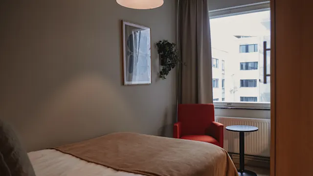En säng med en brun filt placerad i ett litet rum. En röd stol och ett litet bord står vid ett stort fönster som släpper in naturligt ljus. Väggkonst och en taklampa fullbordar miljön.