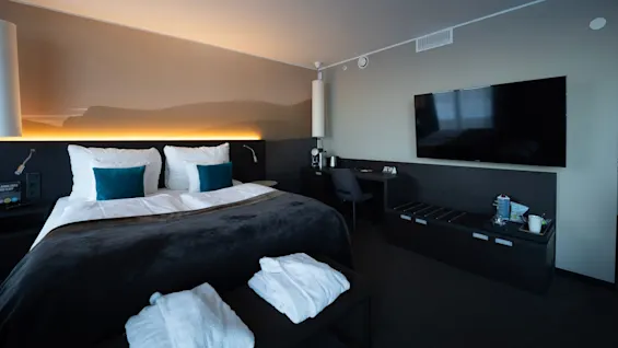 Et pænt indrettet hotelværelse har en dobbeltseng med hvide puder og turkise puder, ved siden af et TV og et skrivebord. To badekåber ligger på en nærliggende bænk.