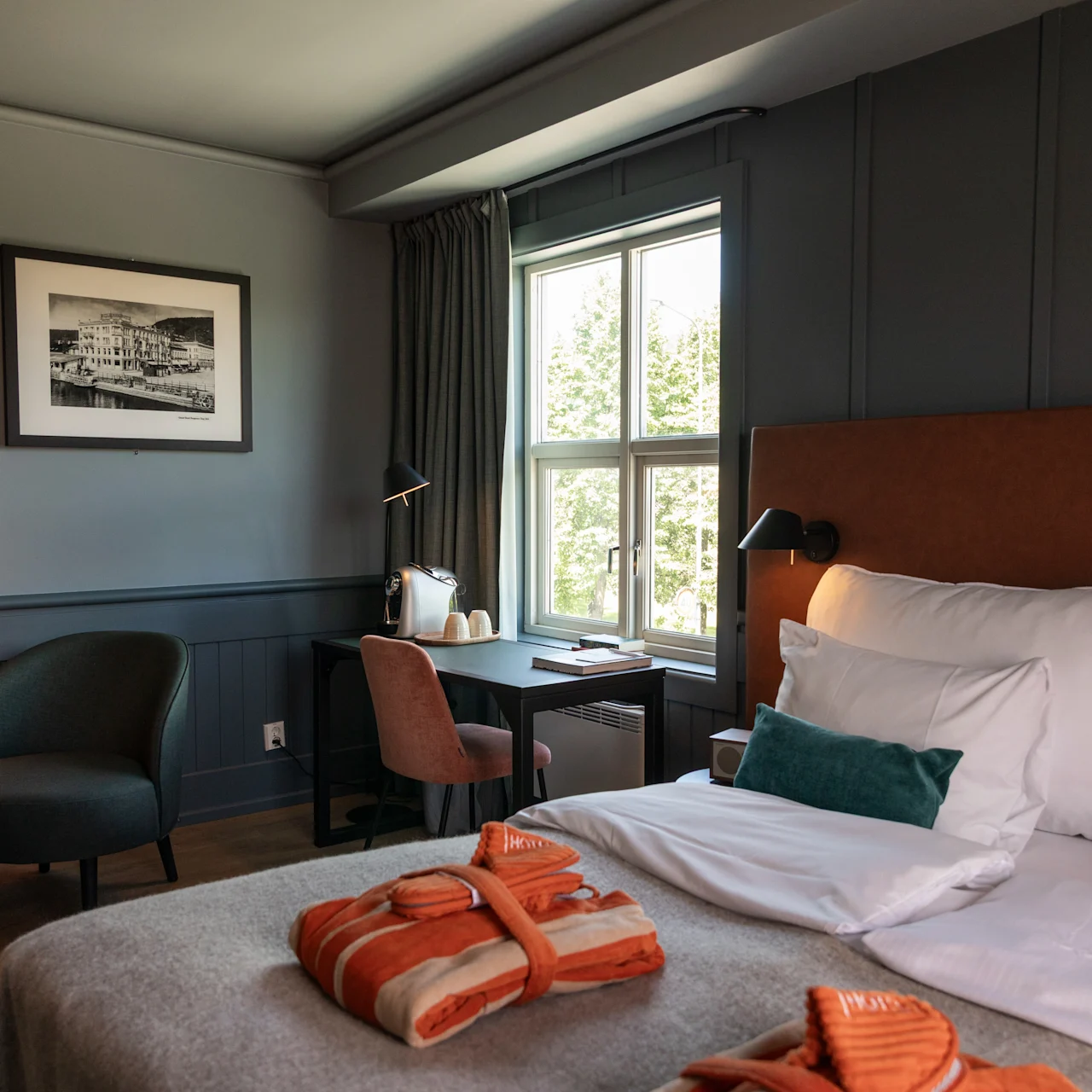 Et pænt arrangeret hotelværelse har en seng med hvidt sengetøj og orange badekåber, ved siden af et vindue. Et skrivebord med lampe og stol giver arbejdsplads ved siden af sengen.