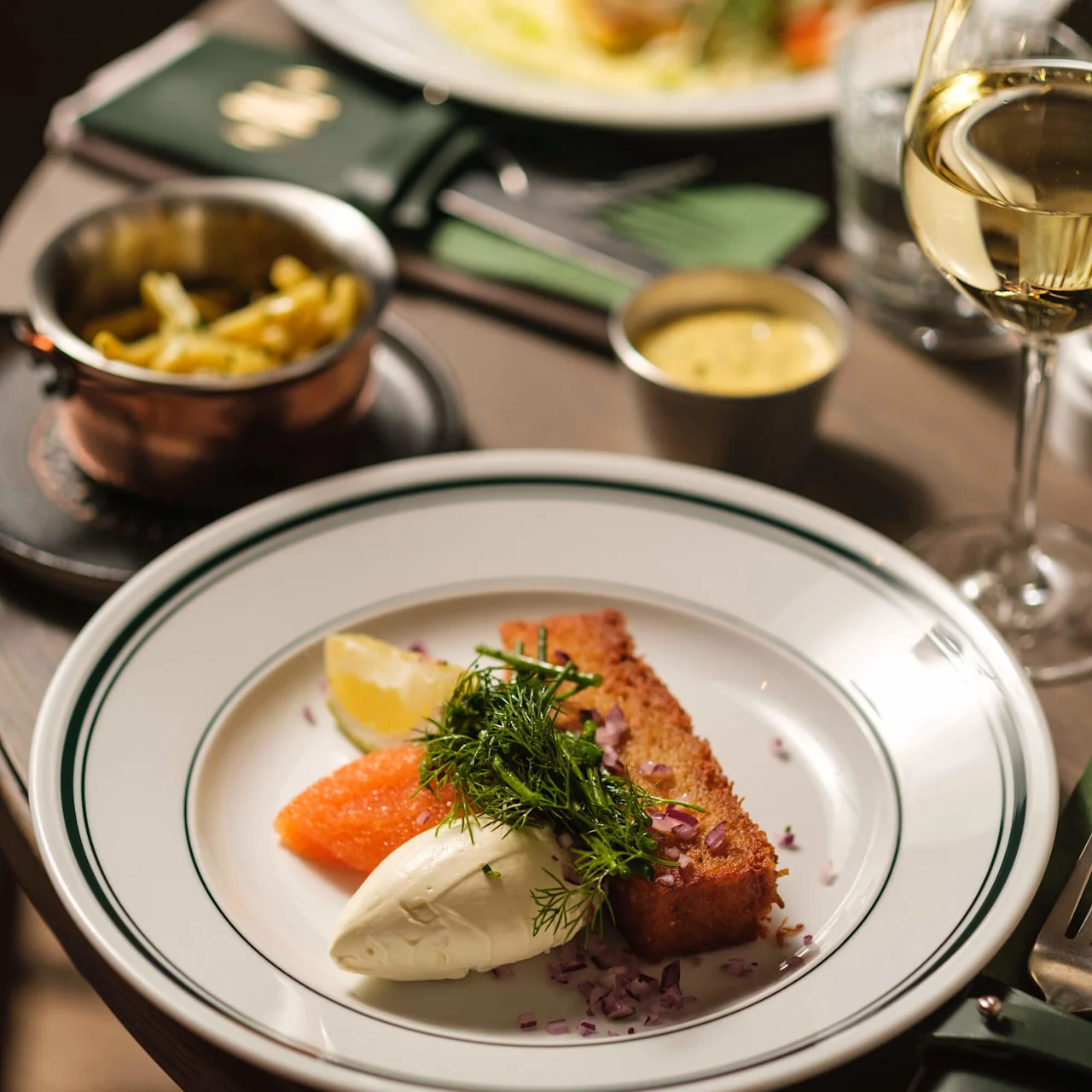 Tallrik med rostat bröd, grädde, laxrom och färsk dill. Serveras på ett bord med bestick, vinglas, en sidorätt och en grön servett.
