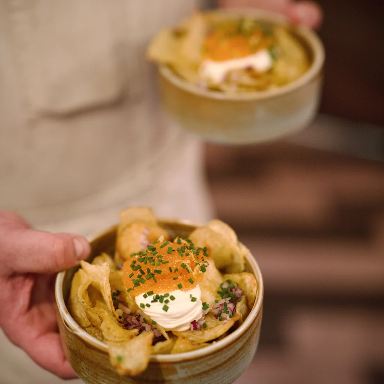 En hånd holder to keramiske skåle med sprøde chips toppet med creme fraiche, purløg og orange garniture, mod en sløret køkkenbaggrund.