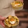 En hand håller två keramiska skålar med krispiga chips toppade med gräddfil, gräslök och orange garnering mot en suddig köksbakgrund.