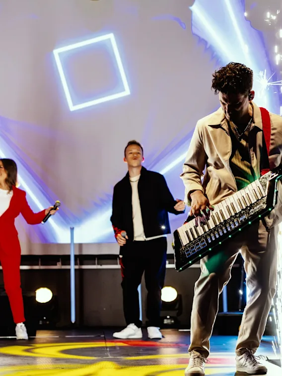 Live band on stage with singers, guitar player, and person in front of image playing a keytar, with sparks on stage