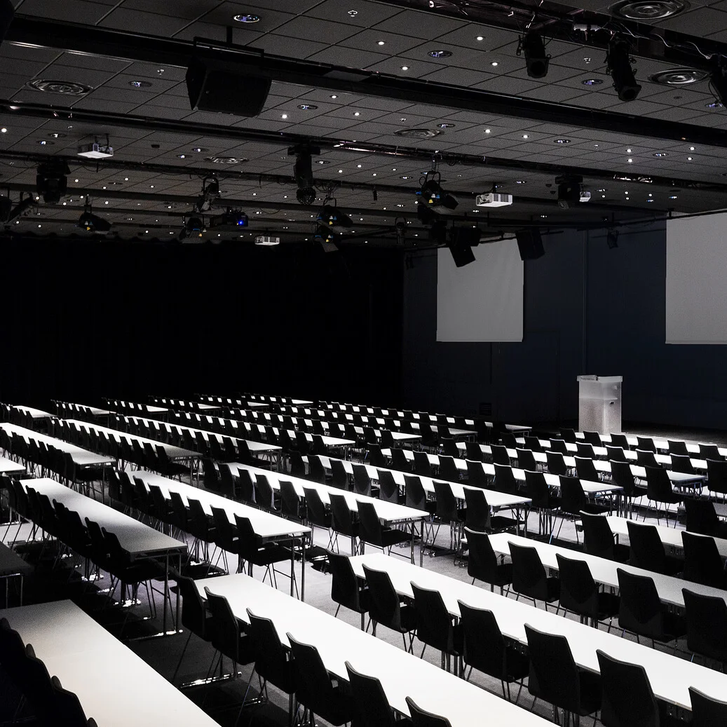 An empty conference room with rows of white tables and black chairs, facing a stage with a podium. The room is dimly lit with projectors on the ceiling.
