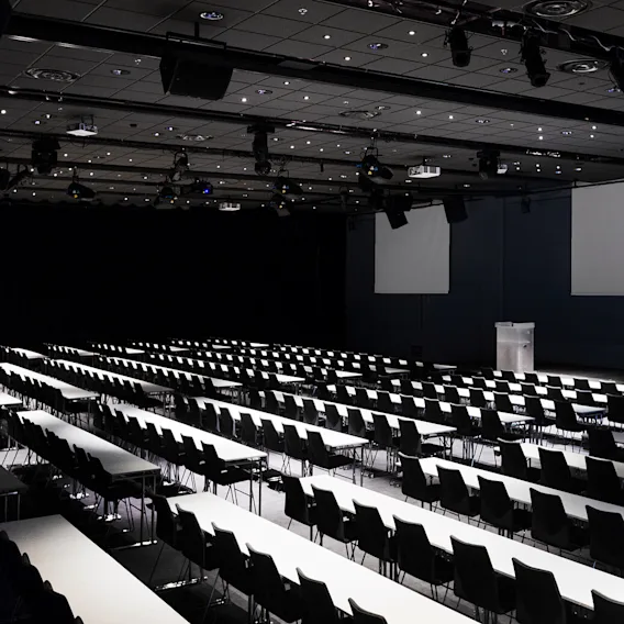 En tom konferansesal med rader av hvite bord og svarte stoler, vendt mot en scene med en talerstol. Rommet er svakt opplyst med projektorer i taket.