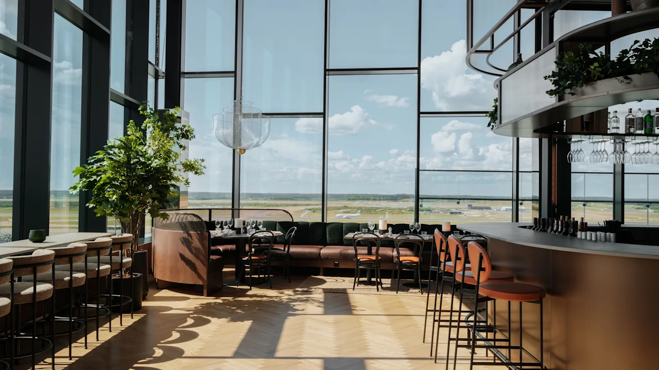 En moderne restaurant og bar med store vinduer som vender ut mot en flyplassrullebane, med stilige sitteplasser og en velfylt bar.