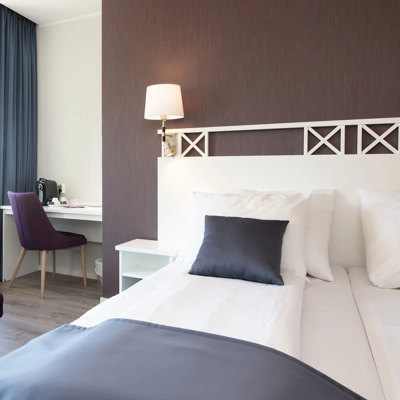 Ett prydligt hotellrum med en plyschlila fåtölj, vit sängklädsel och ett arbetsbord.
