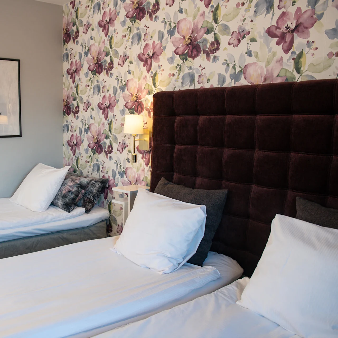 Two neatly made single beds stand in a hotel room with floral wallpaper, a padded headboard, and framed sailboat photographs. A wall lamp softly illuminates the space.