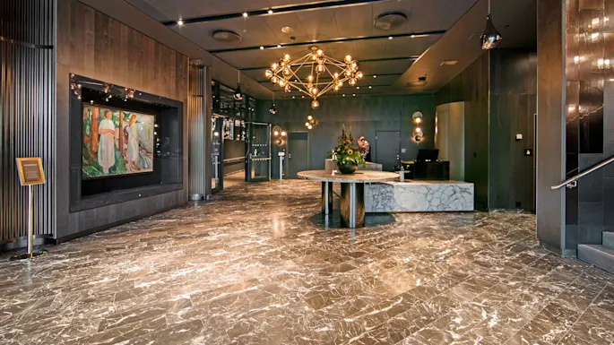 Elegant chandelier hangs above a marble table with flowers in a luxurious lobby. Modern artwork decorates the dark wood wall, enhancing the sophisticated ambiance. Text: none.