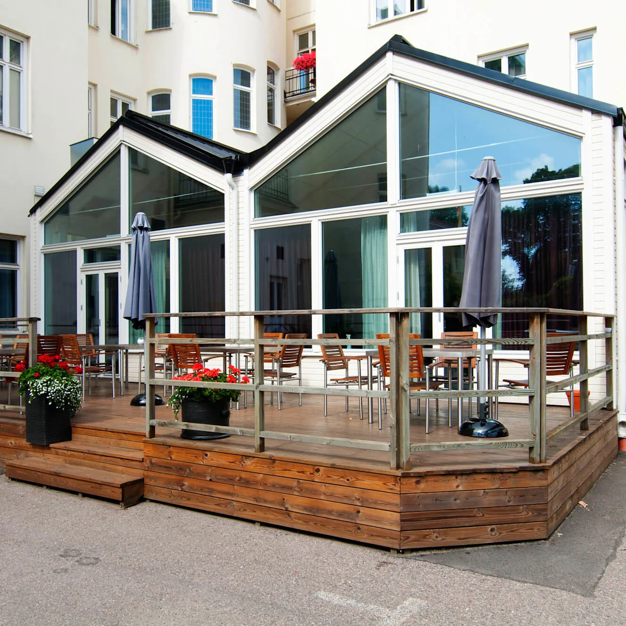 A glass-enclosed patio with wooden decking and outdoor seating, outside a white building.