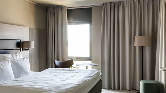 Bed with white linens placed against the left wall; light spills through a window, surrounded by beige curtains. A chair, small table, and floor lamp occupy the room.