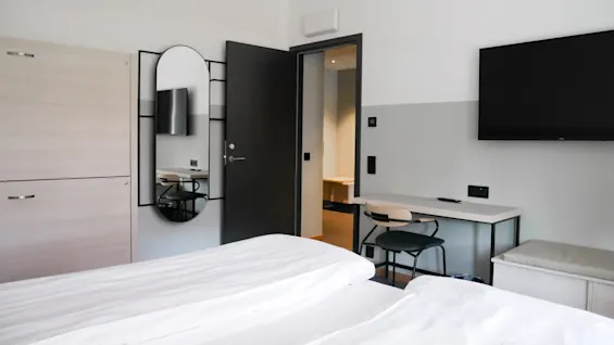 Et minimalistisk hotellrom har en pent oppredd seng. Et speil og en garderobe er til venstre, et skrivebord og en stol står mot høyre vegg, og en TV henger over.