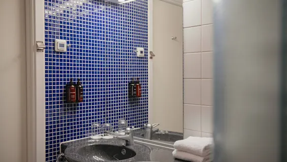 A well-lit bathroom at Home Hotel Uppsala with a dark counter, sink, mirror, blue mosaic tiles, and fresh towels.