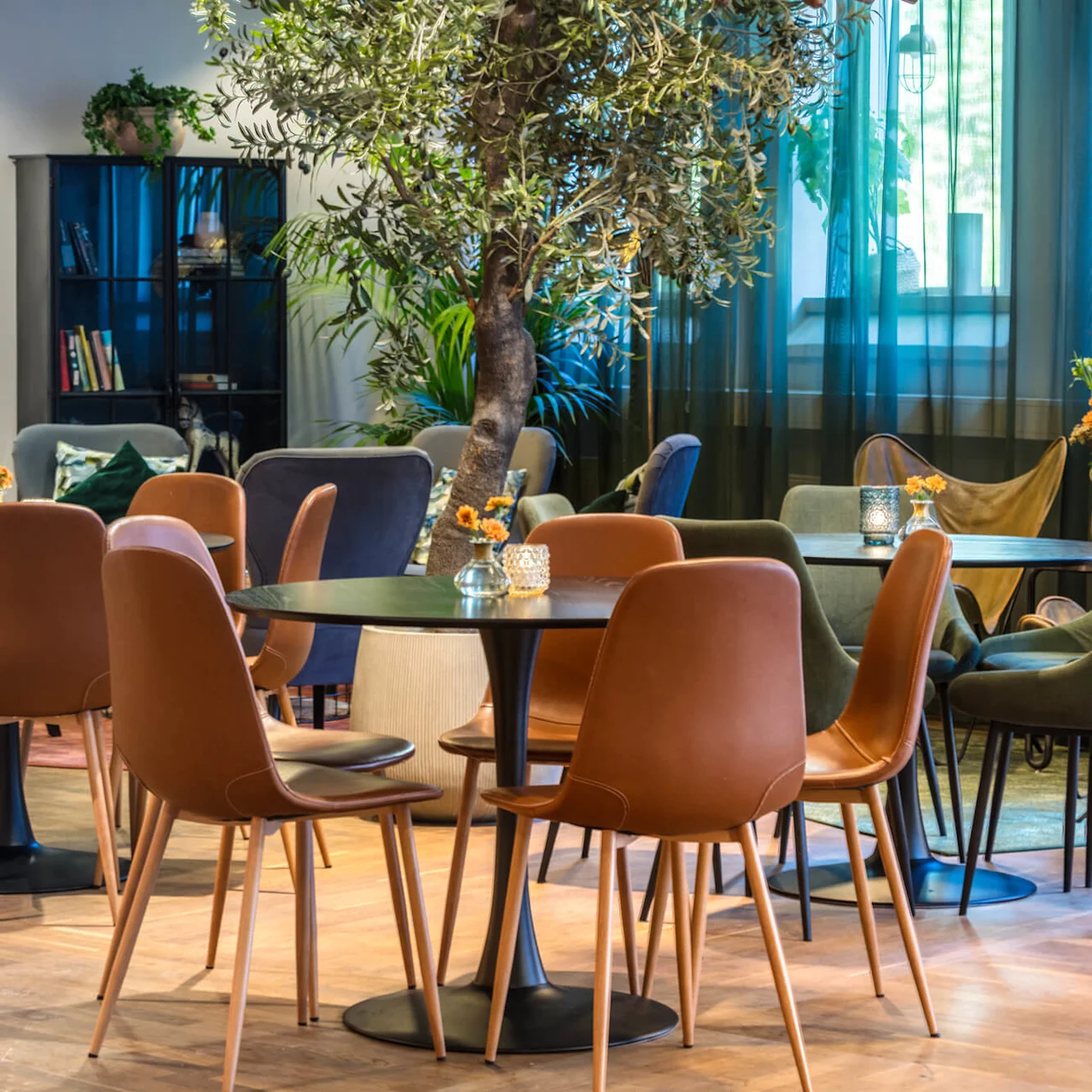 Elegant dining area with round tables and a mix of brown and grey upholstered chairs, amidst indoor plants and modern decor.