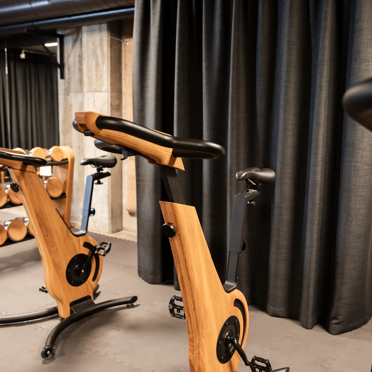 Motionscykler med trærammer står stille i et træningscenter, omgivet af sorte gardiner og et stativ med håndvægte.