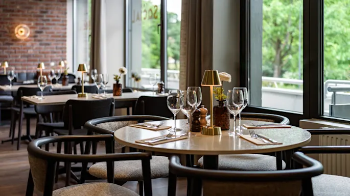 Modern dining table set with wine glasses, cutlery, and a small lamp, in a restaurant with brick walls and large windows overlooking green trees.