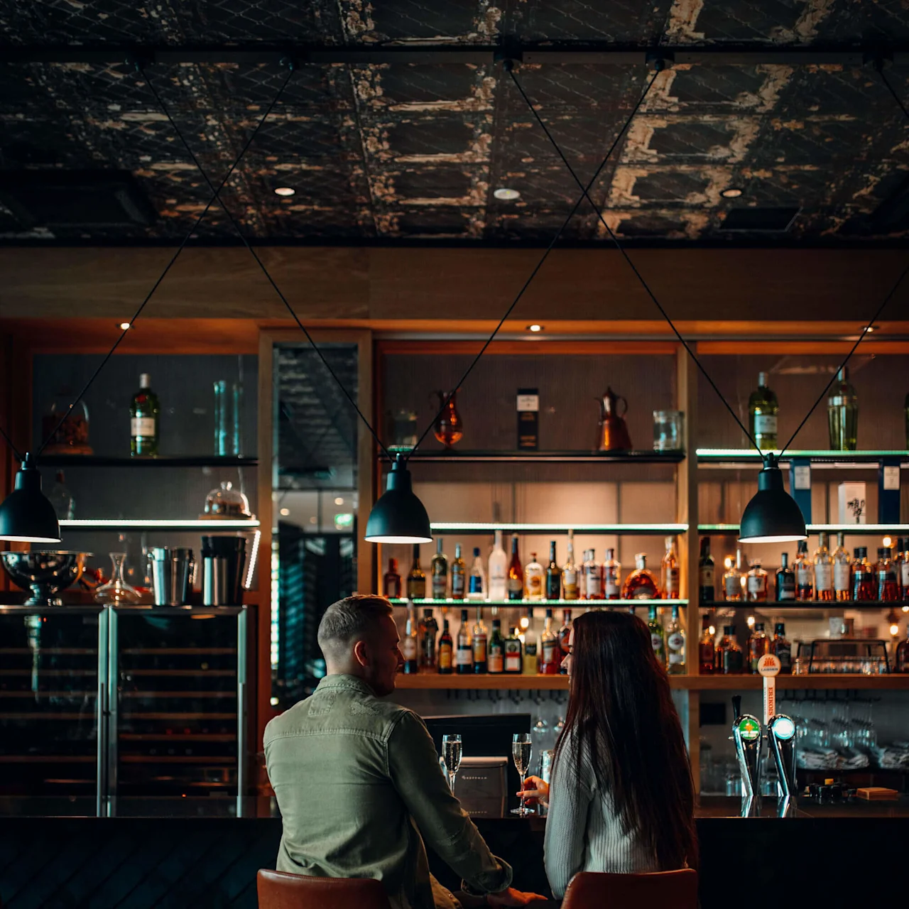 Two people sit on bar stools, talking and holding drinks in a dimly lit bar. Shelves behind them display various bottles under soft lighting.