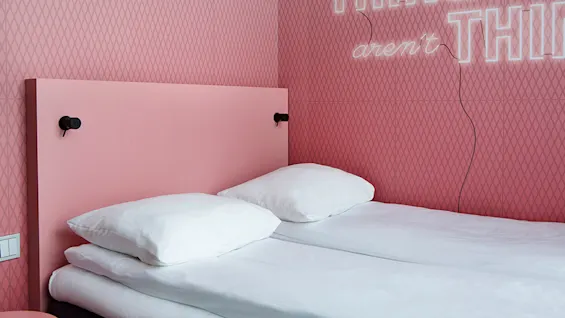 En seng med to hvite puter og et rosa sengegavl er plassert mot en rosa mønstret vegg med neontekst: "aren't THINGS".