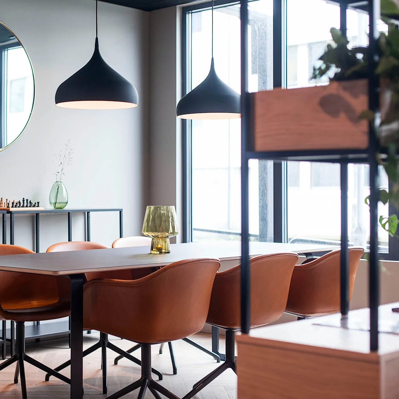 Spisebord omgitt av brune stoler, plassert i et moderne rom med store vinduer. Over henger to svarte pendellamper, mens planter og dekorasjoner pryder hyller mot veggen.