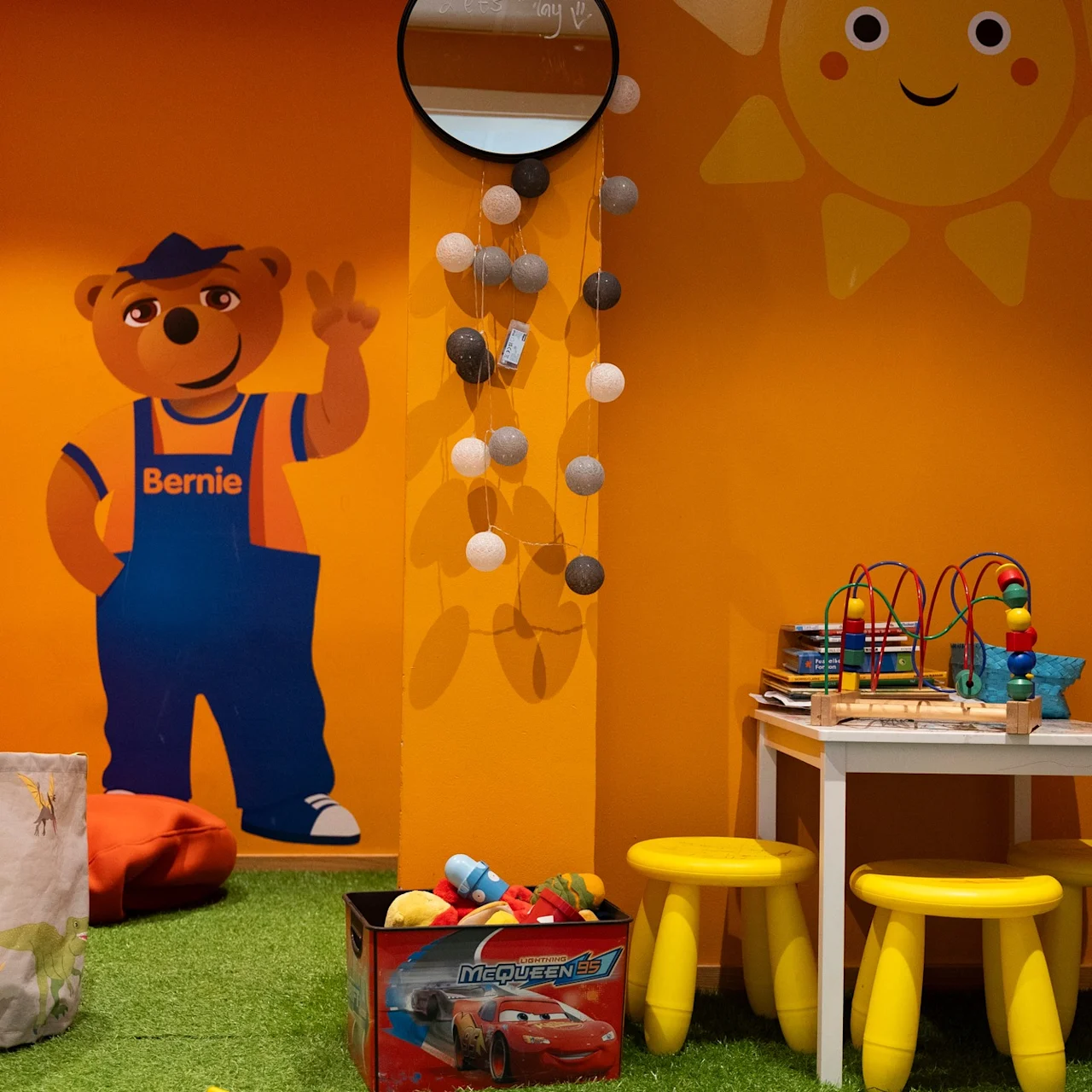 Ett lekrum för barn med en björnmålning kallad "Bernie" på en orange vägg, ett litet bord med pallar och leksaker, samt en rad dekorativa bollar ovanför.