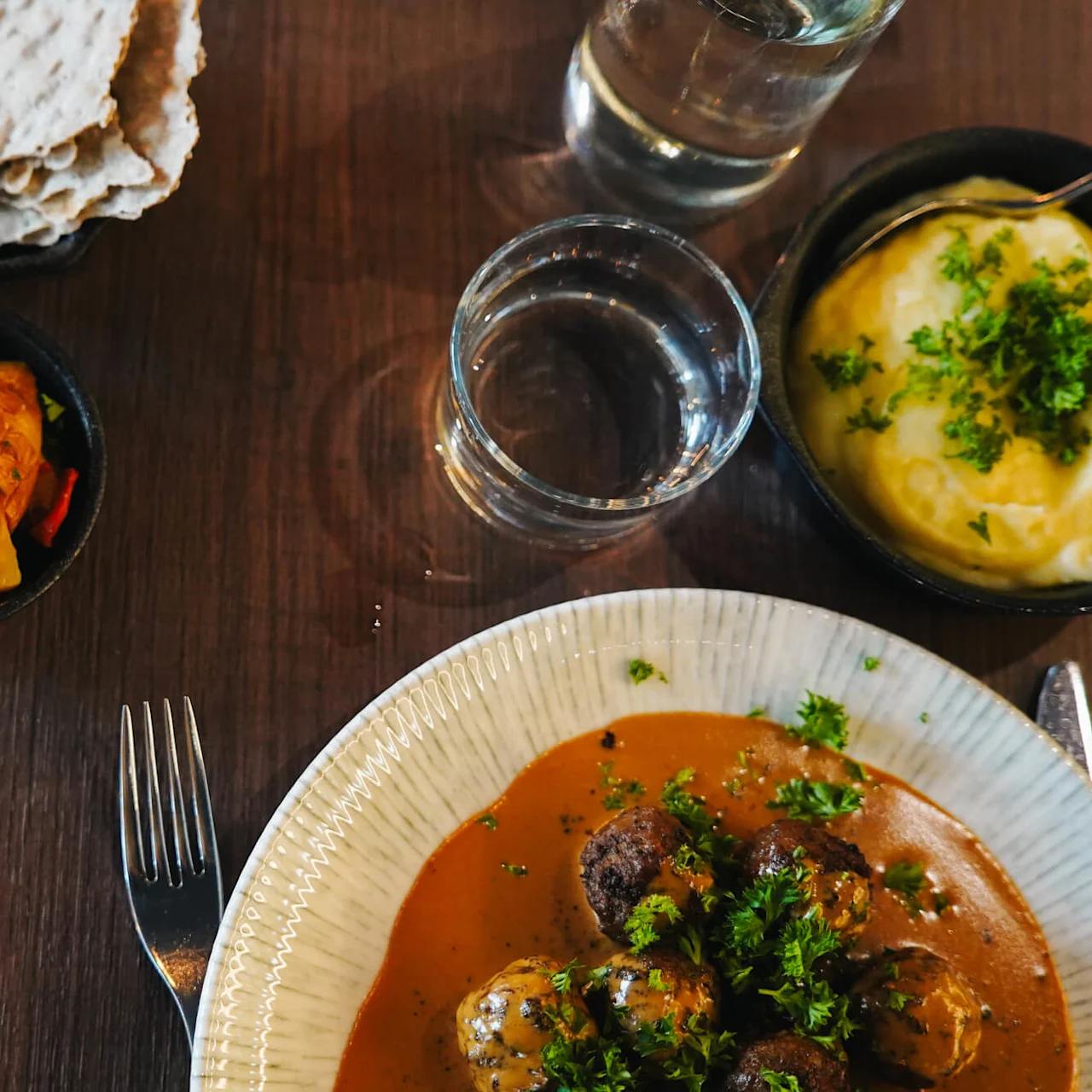 Frikadeller med sovs garneret med persille er på en keramisk tallerken. I nærheden er kartoffelmos, ristede grøntsager, sprødt brød med sennep, et glas vand og en gaffel på et træbord.